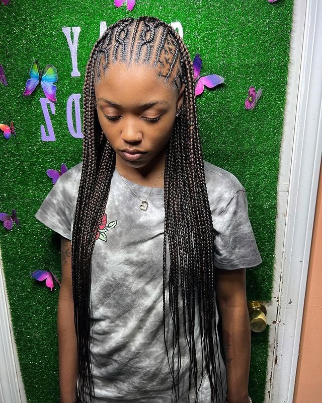 Person with Fulani braids, featuring intricate patterns and adorned with beads, posing against a green leafy background.