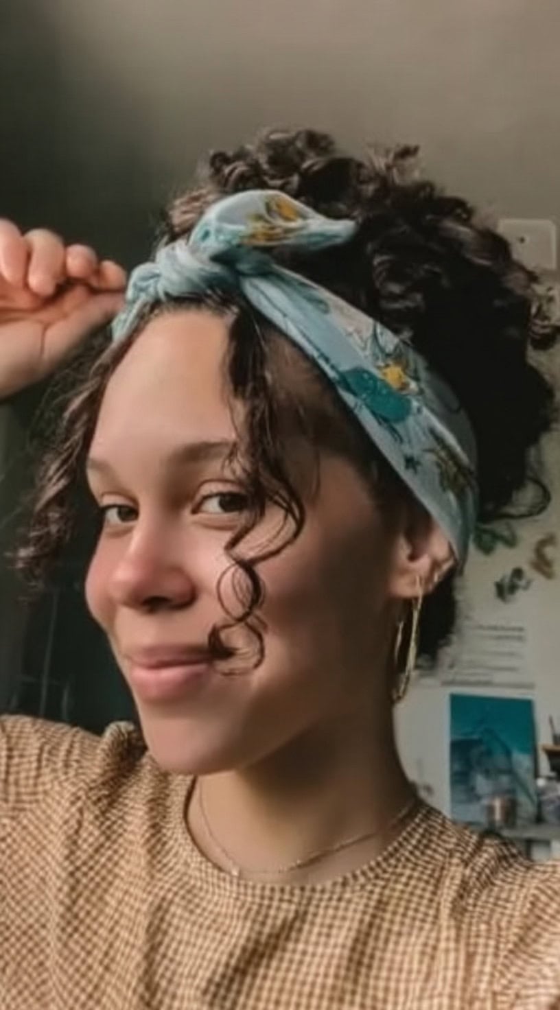 Young woman wearing her 3a curls in a high pineapple updo tied with a floral scarf, with two curls left out in front.