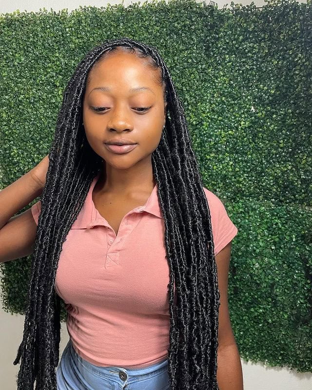 Person with long faux locs wearing a pink polo shirt, posing against a green leafy background.