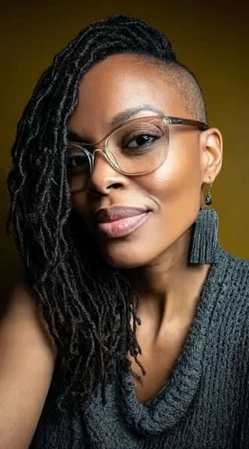 Black woman with long locs swept to one side and a shaved side, wearing glasses.