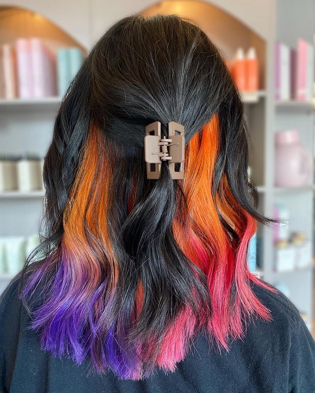 Back view of dark hair with vibrant peekaboo highlights in orange, pink, and purple hues, styled half-up.