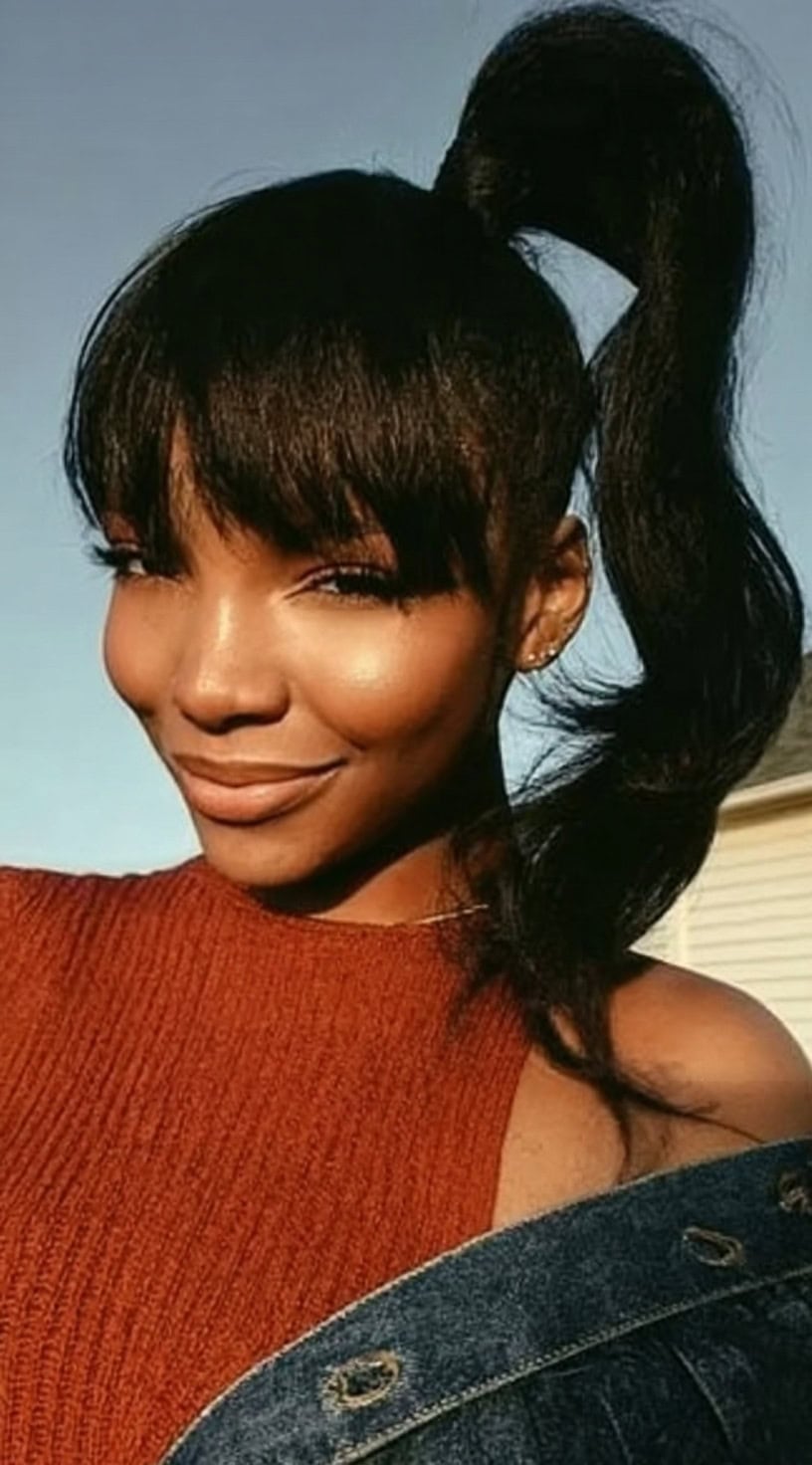 Smiling woman with a fluffy high ponytail and soft full bangs in warm outdoor lighting.