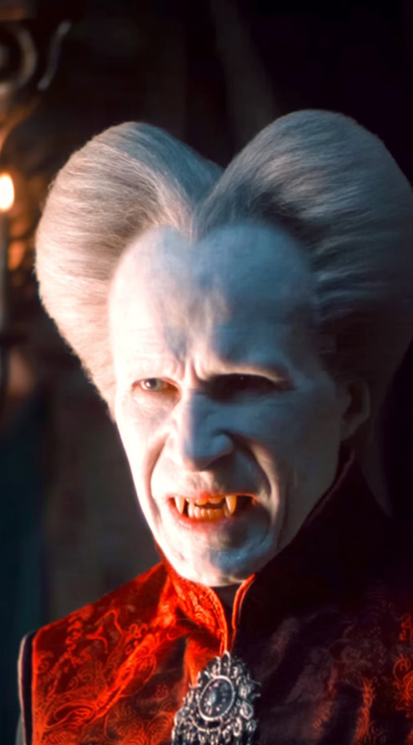 A white-haired vampire with a heart-shaped bouffant hairstyle and embroidered red robe snarls in candlelight.