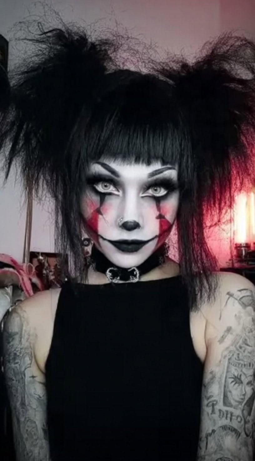 Person with messy, teased black pigtails, blunt bangs, and bold black-and-red clown makeup in an indoor setting.
