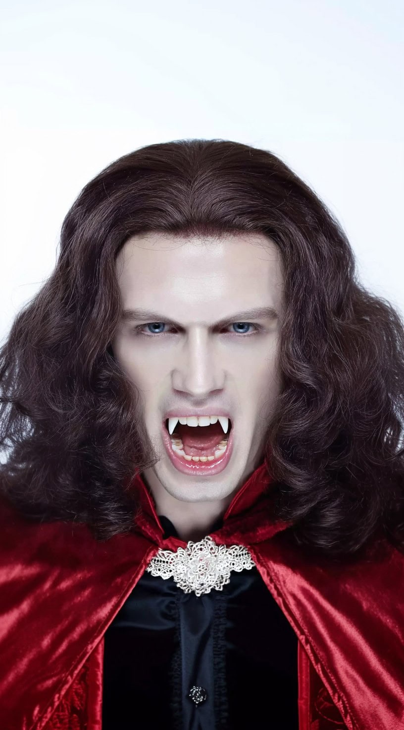 A pale man with voluminous shoulder-length brown hair, parted in the center, snarls with fangs bared while wearing a red cape.