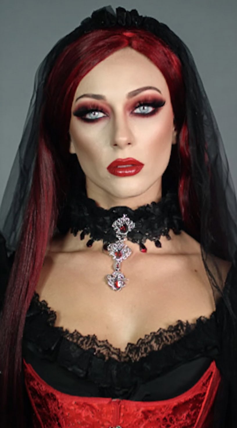 Long, straight dark red hair topped with a black veil and paired with bold makeup.