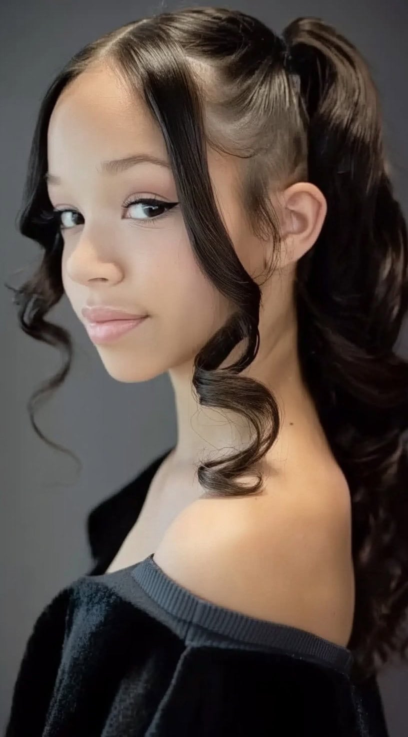 A young woman in a black off-the-shoulder outfit with a sleek high ponytail and two curled face-framing strands.