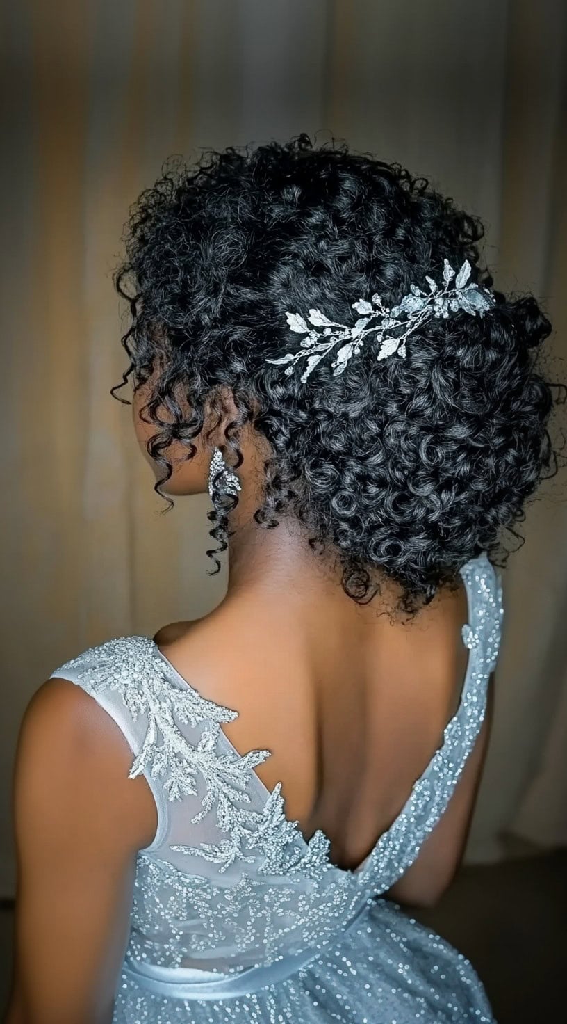 A woman in a shimmering gown with a textured curly updo, adorned with a delicate silver hairpiece at the back.