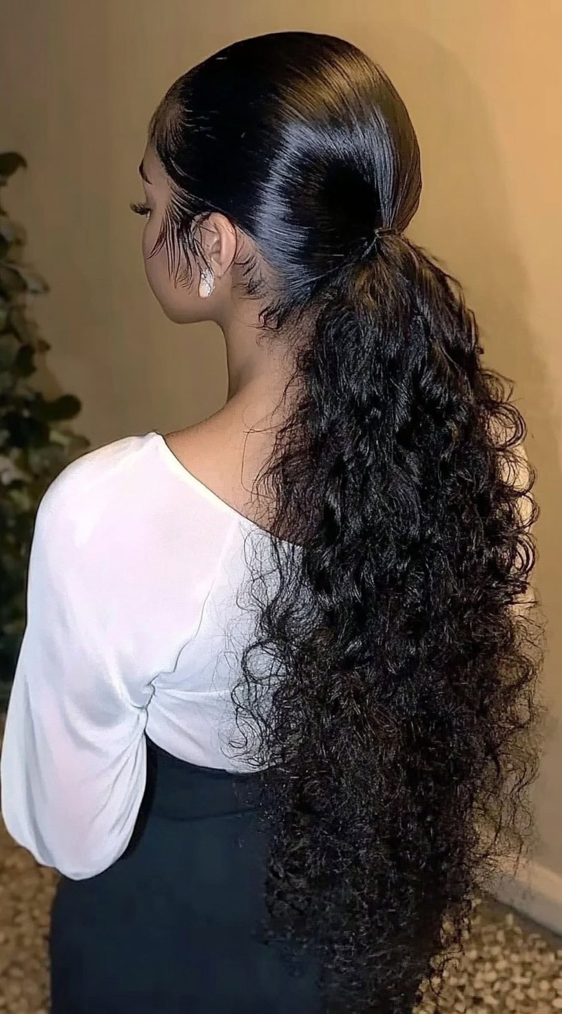 A woman with a sleek, low ponytail transitioning into voluminous natural curls, dressed in a white blouse and black skirt.