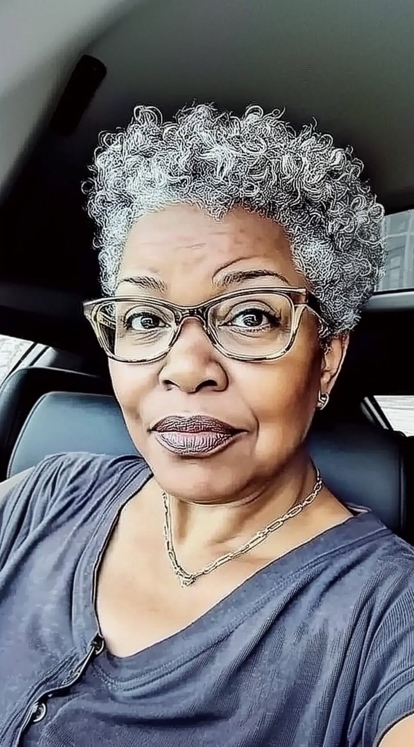 A confident older Black woman with short, voluminous silver curls, wearing glasses and a casual gray top, looking stylish and relaxed.