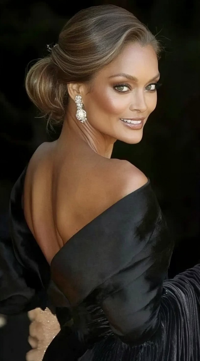 A woman with a sleek, low bun featuring soft waves, complemented by elegant earrings and an off-the-shoulder gown.