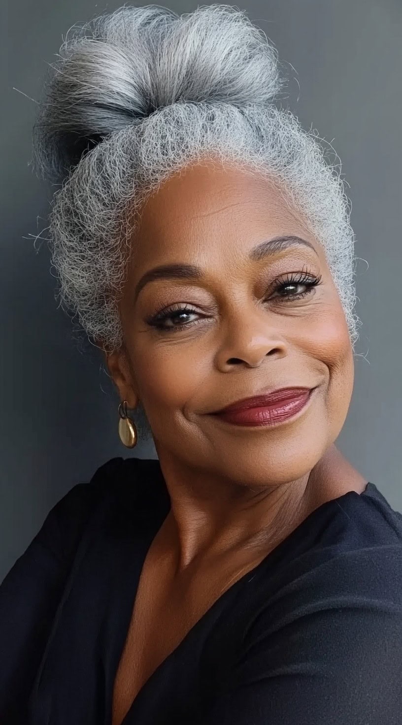 A confident older Black woman with silver-gray natural hair styled into a voluminous high bun, smiling warmly in a black outfit.
