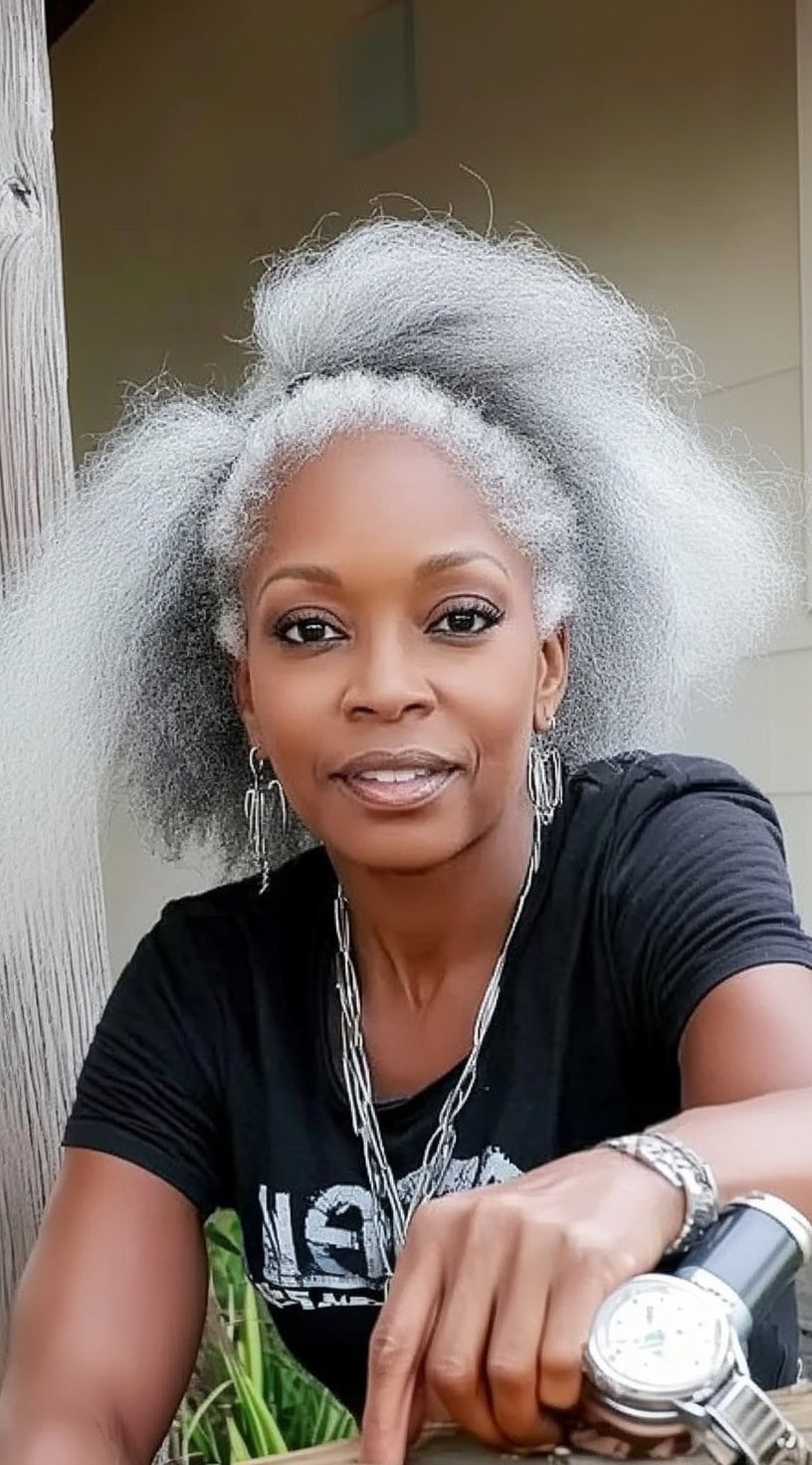 A dynamic older Black woman with a voluminous silver afro, slightly teased for a bold look, wearing a casual black t-shirt and silver accessories.