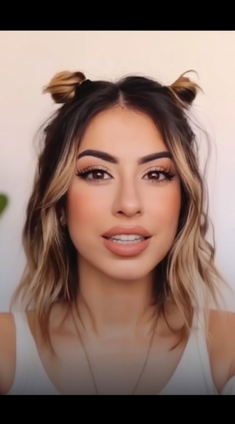 Front view of a woman with chin-length wavy hair styled in two high mini buns, with dark roots and light balayage ends.