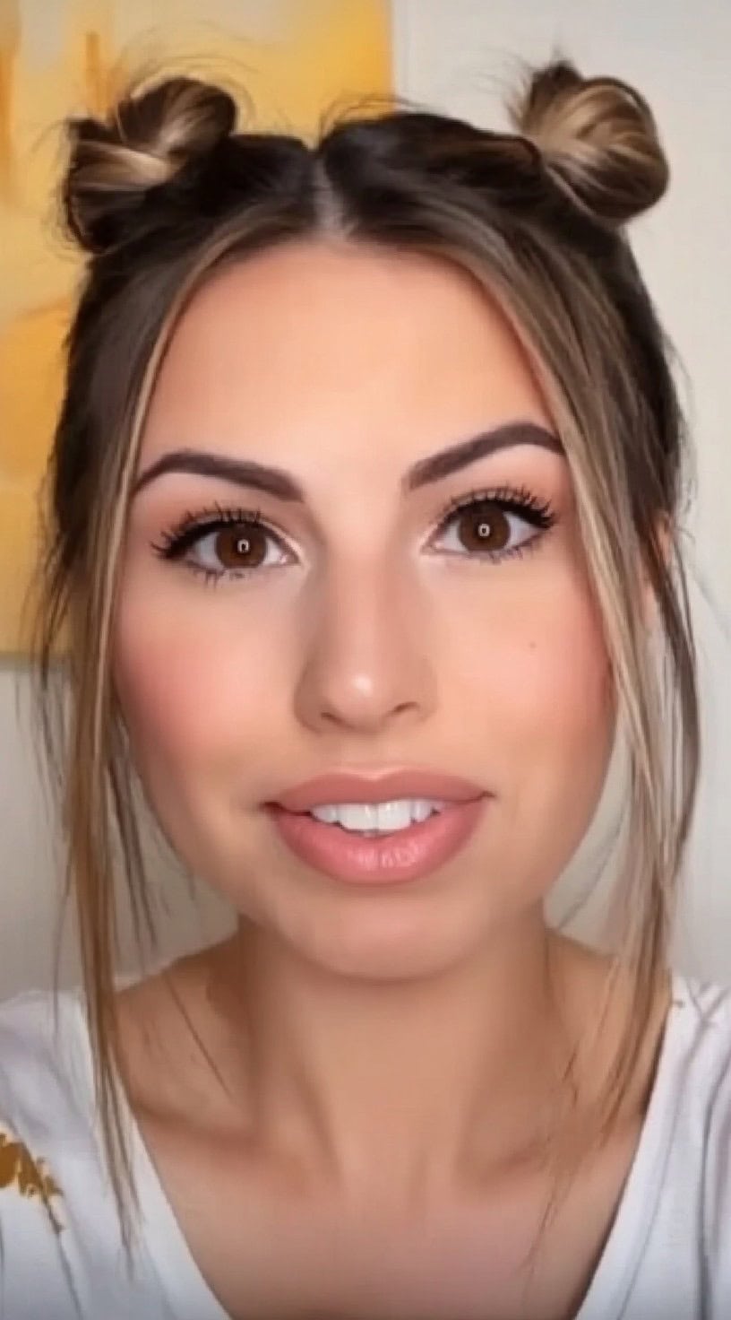 Front view of a woman with two high space buns and soft front tendrils left loose to frame the face.