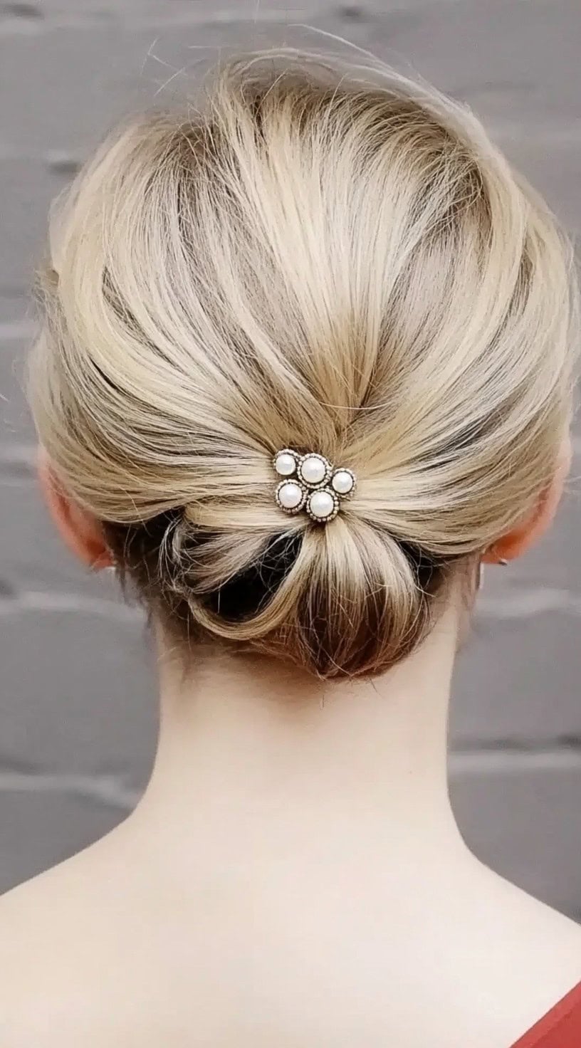 A close-up view of a blonde woman's hair gathered into a low tucked roll, secured with a vintage-style pearl accessory.