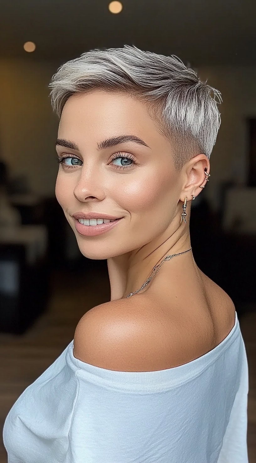 A woman with a frosty silver pixie, featuring volume at the crown and tapered sides, smiling over her shoulder.