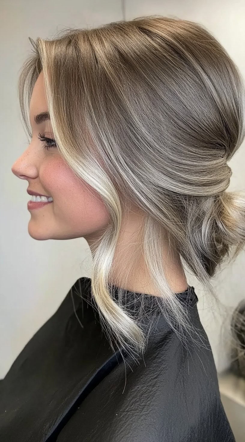 A side view of a woman with a twisted low bun and bold face-framing platinum highlights curled softly around her face.