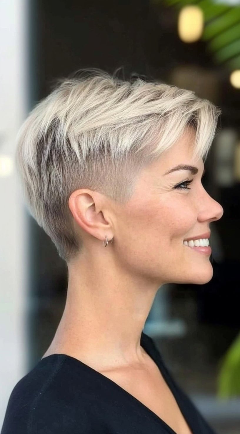 A woman with an icy blonde stacked pixie and long side-swept top layers is smiling in profile.