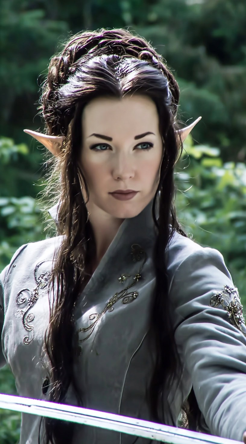 A woman with pointed elf ears and long dark hair styled into a half-up braided crown, wearing a silver embroidered jacket.