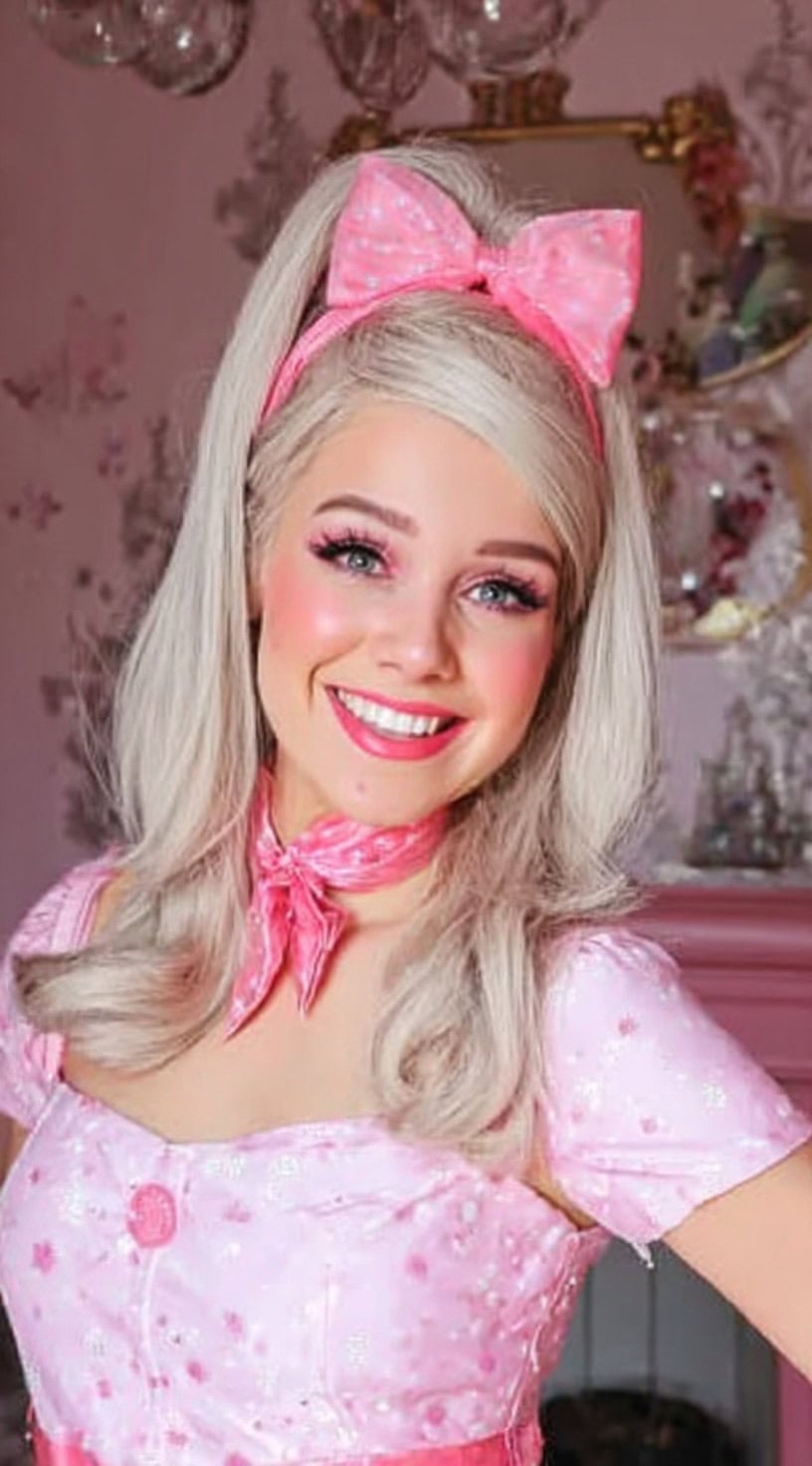 A smiling woman with light blonde hair styled half-up with voluminous flipped ends, topped with a pink polka-dot bow headband and matching scarf.
