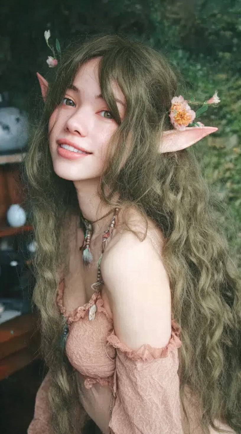 A smiling woman with green-tinted long wavy hair, elf ears, and delicate flowers tucked into her strands.
