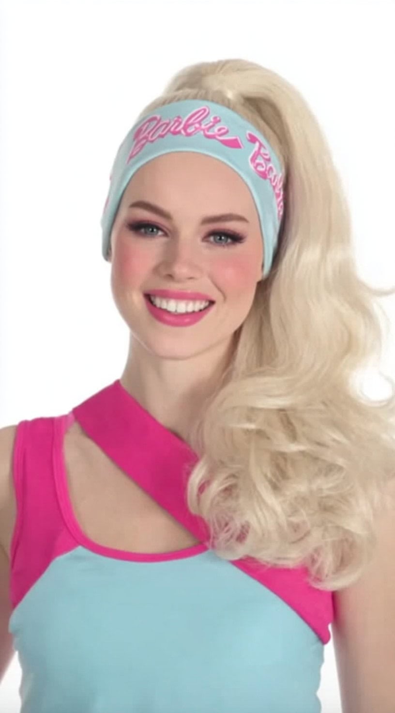 A smiling woman with platinum blonde hair styled in a high curled ponytail with a retro Barbie headband and a color-blocked pink and blue outfit.