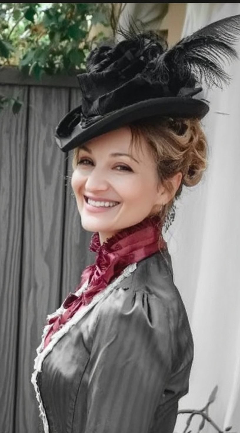 Smiling woman in Victorian attire with a black feathered hat over a softly pinned updo.