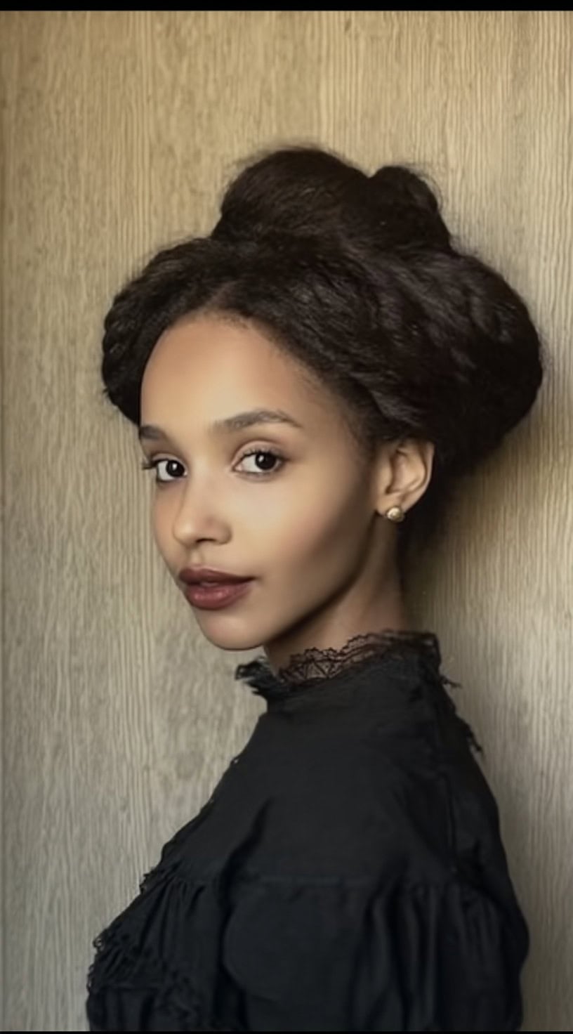 Profile view of a woman with dark hair styled into a large twisted crown bun, paired with a high-collared black dress.